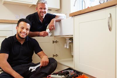 Apprentice plumber working with tradesman under a kitchen sink.