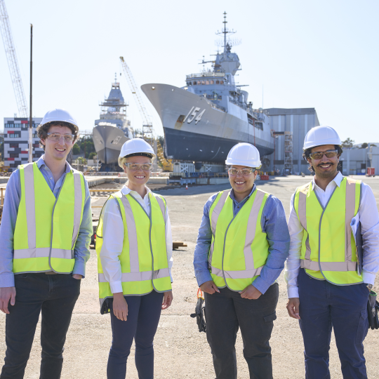 Four defence workers wearing PPE equipment, standing in front of a naval ship 