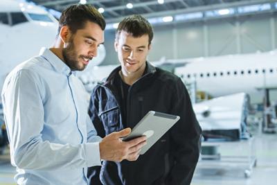 Two male aircraft engineers looking at iPad with planes in a workshop behind them.