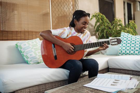 A happy teenager in casual clothing, sitting on a white couch, playing the guitar